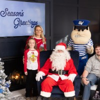 Little girl sits on dad's lap while other sister and mom stand on the side of Louie and Santa